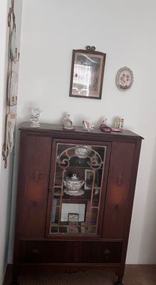 Sale Photo Thumbnail #16: Vintage dark wood china cabinet featuring a central glass display door with decorative fretwork, flanked by carved side panels and a bottom drawer, decorated with porcelain figurines and framed wall art.