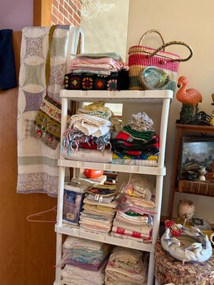 Sale Photo Thumbnail #71: A white plastic shelving unit stacked with vintage linens, crocheted blankets, and quilts, flanked by a woven market bag and a flamingo statuette.