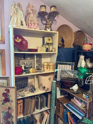 Sale Photo Thumbnail #80: Bookshelves filled with eclectic decor items, including vintage books, angel figurines, framed photos, woven baskets, and an ornate table lamp.