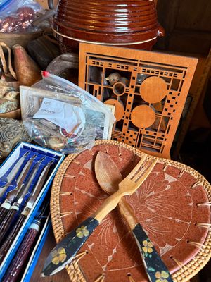 Sale Photo Thumbnail #147: Collection of tabletop decor and kitchenware, featuring Frank Lloyd Wright-style laser-cut wooden trivets, hand-painted wooden serving utensils resting on a leather and wicker tray, and boxed flatware.