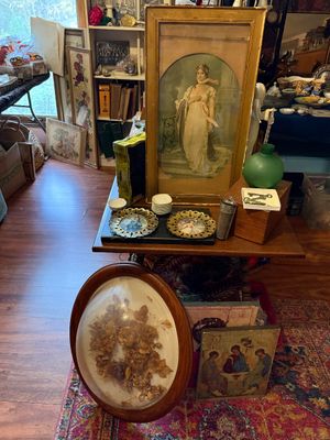 Sale Photo Thumbnail #88: Collection of vintage framed art and home decor, including a large framed portrait print of a woman in a gold frame, an oval convex shadow box with dried flowers, and small ornate decorative items displayed on a wooden table.