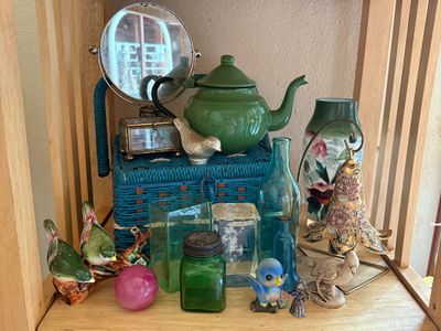 Sale Photo Thumbnail #33: Collection of decorative home accents, including a green enamel teapot, blue woven box, bird figurines, colored glass bottles, and a round vanity mirror displayed on a wooden shelf.