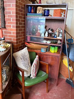 Sale Photo Thumbnail #46: Mid-century wooden hutch displaying various glassware and collectibles, paired with a green vinyl armchair holding decorative pillows.