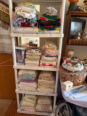 Sale Photo Thumbnail #68: White plastic four-tier shelving unit stacked with vintage linens, crocheted blankets, embroidered cloths, and bed skirts.