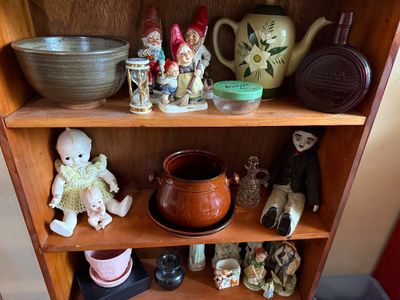 Sale Photo Thumbnail #10: Wooden bookcase filled with vintage collectibles, including ceramic garden gnomes, Kewpie-style dolls, studio pottery, and a Charlie Chaplin figure.