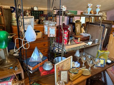 Sale Photo Thumbnail #84: Collection of tabletop decor and collectibles including a green banker's lamp, white essential oil diffuser, jarred candles, framed art, and vintage glassware arranged on shelving.