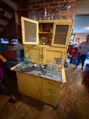 Sale Photo Thumbnail #113: Vintage painted yellow Hoosier-style kitchen cabinet featuring glass-front upper doors, zinc work surface, and lower drawers, display-stacked with assorted glassware and vintage kitchenware.