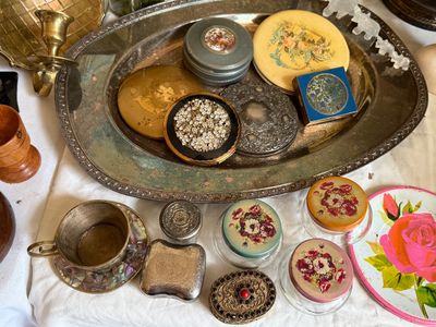 Sale Photo Thumbnail #22: Collection of vintage vanity items and compacts arranged on a silver-plated oval tray alongside trinket boxes, glass jars with floral lids, and small metal decor.