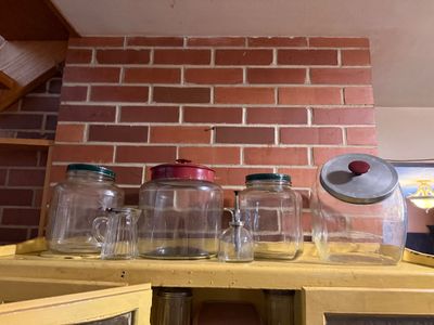 Sale Photo Thumbnail #114: Collection of vintage glass jars, canisters, and a syrup dispenser lined up on top of a yellow cabinet against a brick wall.