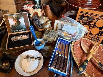Sale Photo Thumbnail #148: Eclectic collection of vintage collectibles, including a John Wayne commemorative pocket knife in a wooden box, stag-handle steak knives, enamelware dishes, and decorative wood carvings.