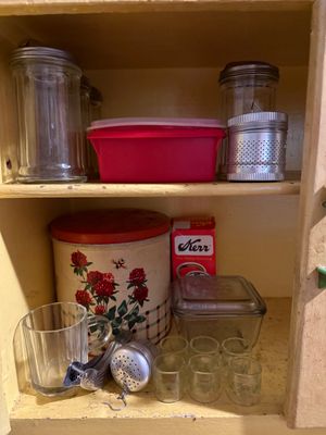 Sale Photo Thumbnail #115: Vintage kitchen cabinet contents including a floral tin canister, Kerr canning jar lids, glass salt shakers, a tea infuser, and small glass shot glasses.