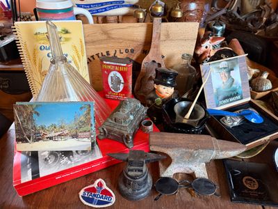 Sale Photo Thumbnail #150: Collection of vintage collectibles including miniature anvils, a Prince Albert tobacco tin, John Wayne memorabilia, glass decanter, postcards, and small figurines.