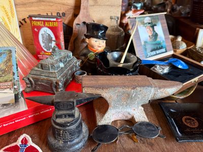 Sale Photo Thumbnail #149: Collection of vintage collectibles including small cast iron anvils, a Prince Albert tobacco tin, a John Wayne portrait box, and round spectacles.