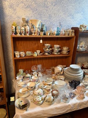 Sale Photo Thumbnail #100: Collection of vintage porcelain and glassware, including floral teacups, saucers, decorative eggs on pedestals, pitchered creamers, and fine china dinnerware displayed on a table and wooden shelving unit.