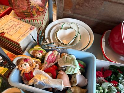 Sale Photo Thumbnail #122: Selection of vintage Christmas ornaments and ceramic figurines, including angels and bonneted girls, displayed in a blue bin alongside white ceramic dishes and vintage cookbooks.