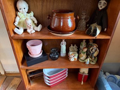 Sale Photo Thumbnail #9: Wooden shelving unit displaying vintage porcelain dolls, figurines, a brown stoneware pot, a pink planter, and red loaf pans.