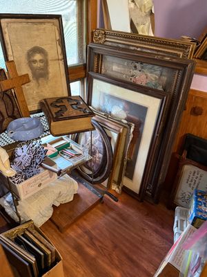 Sale Photo Thumbnail #93: Collection of framed vintage wall art, including a portrait of Jesus Christ, an ornate leather-bound Bible with embossed cross, and various wooden picture frames stacked against a wall.