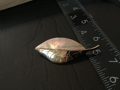 Sale Photo Thumbnail #72: A vintage Liz Claiborne leaf brooch featuring a gold-tone metal half with detailed veining and a iridescent mother-of-pearl (or shell inlay) half. Signed 'LC' on the back. Measures approximately 2.25 inches long. Features a standard rollover safety clasp 