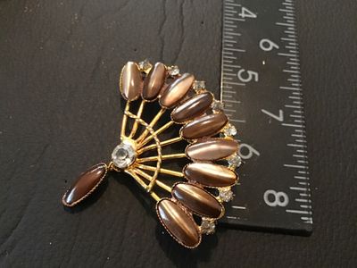 Sale Photo Thumbnail #79: A vintage fan-shaped costume jewelry brooch featuring gold-tone metal, brown marquise/cabochon stones with a cat's eye sheen, clear rhinestone accents along the outer rim, and a dangling accent piece. Measures approximately 2.5 inches across. Shows minor 