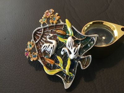 Sale Photo Thumbnail #88: Silver-tone bell-shaped brooch pin featuring colorful enamel artwork depicting two white swans in a pond setting with cattails and floral accents. Translucent stained glass-style back in shades of teal, orange, and blue. Measures approximately 2 inches wi