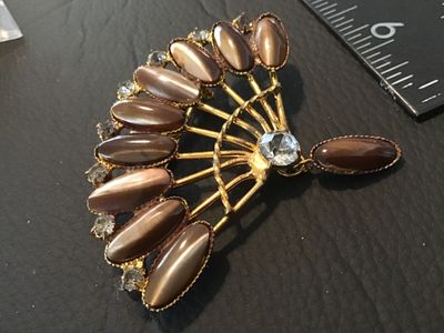 Sale Photo Thumbnail #78: A vintage fan-shaped costume jewelry brooch featuring gold-tone metal, brown marquise/cabochon stones with a cat's eye sheen, clear rhinestone accents along the outer rim, and a dangling accent piece. Measures approximately 2.5 inches across. Shows minor 