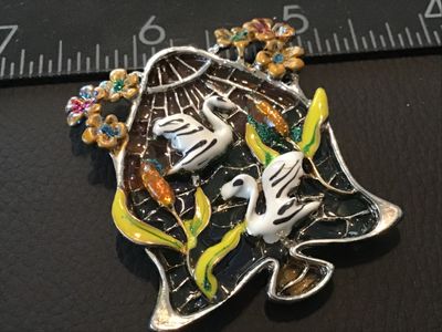 Sale Photo Thumbnail #90: Silver-tone bell-shaped brooch pin featuring colorful enamel artwork depicting two white swans in a pond setting with cattails and floral accents. Translucent stained glass-style back in shades of teal, orange, and blue. Measures approximately 2 inches wi