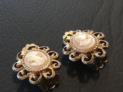 Sale Photo Thumbnail #86: Pair of vintage clip-on earrings featuring Shell cameo portrait  set within ornate, filigree-style gold-tone metal frames. The clips show some age-related patina and finish wear on the reverse side. Overall good vintage condition.