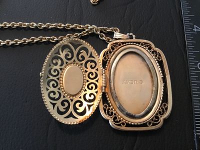 Sale Photo Thumbnail #11: Vintage Avon gold tone filigree locket necklace featuring a blue faux lapis lazuli cabochon stone on the front cover. The locket opens on a side hinge to reveal space inside, marked AVON ©. Pendant measures approximately 2 inches in length. Comes attache