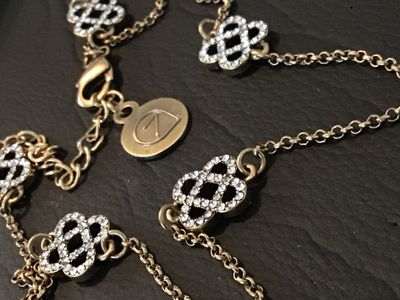 Sale Photo Thumbnail #65: Gold-tone chain necklace featuring openwork, Celtic knot-style stations encrusted with clear rhinestones. Includes a lobster claw clasp with an adjustable extender chain and a signature round logo hangtag with a stylized '7' or leaf design. Gently used co