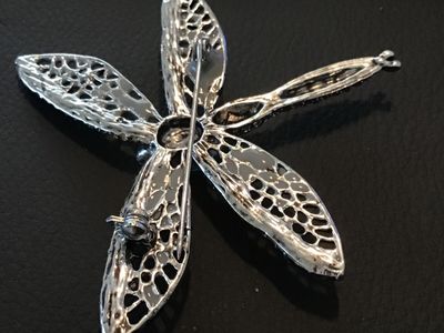 Sale Photo Thumbnail #95: Silver-tone dragonfly brooch featuring intricate openwork filigree wings, dark blue and clear accent rhinestones, and a large bright blue center stone. Includes a standard pin back with a safety clasp. Measures approximately 2.5 inches long. In good condi