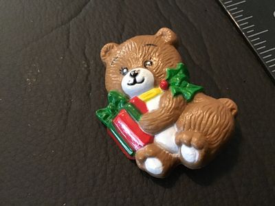 Sale Photo Thumbnail #55: Vintage Christmas-themed teddy bear pin or brooch by Russ. Made of molded painted plastic, featuring a teddy bear holding a wrapped present with holly leaf details. Marked 'Russ' and 'Made in Hong Kong' on the back. Pin mechanism is intact and functional.
