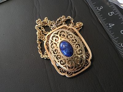 Sale Photo Thumbnail #8: Vintage Avon gold tone filigree locket necklace featuring a blue faux lapis lazuli cabochon stone on the front cover. The locket opens on a side hinge to reveal space inside, marked AVON ©. Pendant measures approximately 2 inches in length. Comes attache