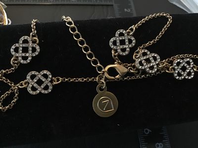 Sale Photo Thumbnail #66: Gold-tone chain necklace featuring openwork, Celtic knot-style stations encrusted with clear rhinestones. Includes a lobster claw clasp with an adjustable extender chain and a signature round logo hangtag with a stylized '7' or leaf design. Gently used co
