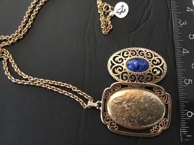 Sale Photo Thumbnail #10: Vintage Avon gold tone filigree locket necklace featuring a blue faux lapis lazuli cabochon stone on the front cover. The locket opens on a side hinge to reveal space inside, marked AVON ©. Pendant measures approximately 2 inches in length. Comes attache