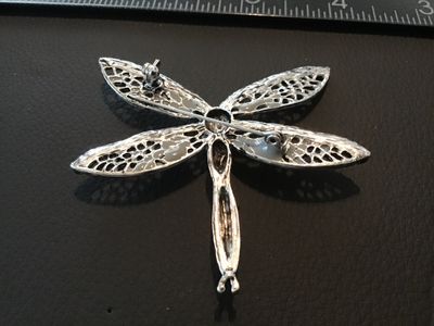 Sale Photo Thumbnail #94: Silver-tone dragonfly brooch featuring intricate openwork filigree wings, dark blue and clear accent rhinestones, and a large bright blue center stone. Includes a standard pin back with a safety clasp. Measures approximately 2.5 inches long. In good condi