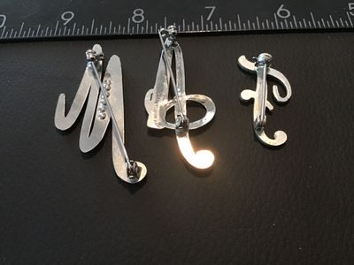 Sale Photo Thumbnail #112: Set of three silver-tone initial letter brooches featuring the letters 'M', 'A', and 'F'. Each brooch is set with clear rhinestones in a script design with pin-back closures. Brooches measure approximately 1.25 to 2.25 inches in height. Items show light w