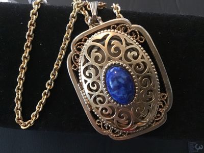 Sale Photo Thumbnail #7: Vintage Avon gold tone filigree locket necklace featuring a blue faux lapis lazuli cabochon stone on the front cover. The locket opens on a side hinge to reveal space inside, marked AVON ©. Pendant measures approximately 2 inches in length. Comes attache