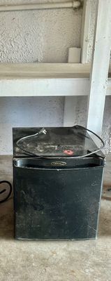 Sale Photo Thumbnail #4: Black Danby Designer compact mini refrigerator showing surface dust and light cosmetic wear, sitting on a concrete floor beneath shelving.