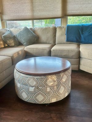 Sale Photo Thumbnail #8: Round upholstered storage ottoman with a wood tray top, set in front of a beige sectional sofa with accent pillows.