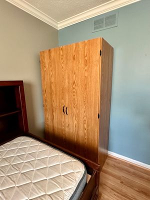 Sale Photo Thumbnail #16: Tall light oak finish laminate armoire or storage cabinet with black double-door handles, positioned in the corner of a bedroom next to a dark wood bed frame.