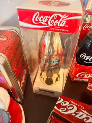 Sale Photo Thumbnail #16: Vintage Coca-Cola Atlanta 1996 Centennial Olympic Games commemorative gold-tone contour bottle, housed in its original clear display box alongside other Coca-Cola memorabilia.