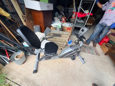 Sale Photo Thumbnail #69: Exerpeutic recumbent stationary exercise bike with digital display monitor, black padded seat, and silver frame, shown in a crowded garage setting.