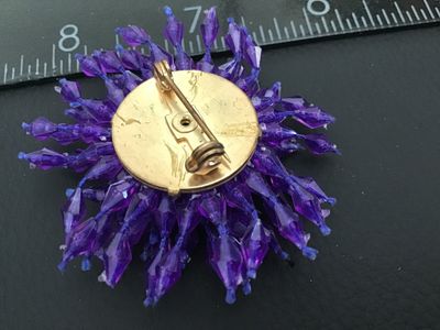 Sale Photo Thumbnail #268: A vintage starburst or pom-pom style brooch featuring faceted purple plastic beads wired/strung onto a gold-tone metal base with a rollover safety pin clasp. Measures approximately 2 inches across. Good pre-owned condition with light vintage wear to the m