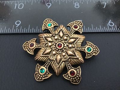 Sale Photo Thumbnail #146: Vintage ornate star / Maltese cross brooch crafted in a textured antique gold-tone metal. Detailed with raised beaded accents, geometric star patterning, and set with red and green rhinestones. Features a standard roll-over safety clasp pin back. Measures