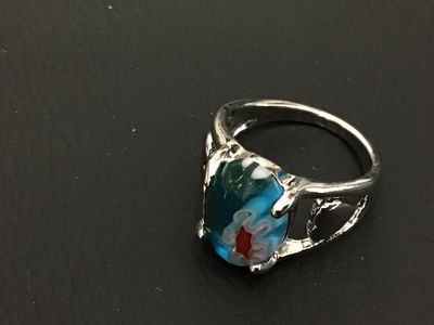Sale Photo Thumbnail #163: Silver-tone metal statement ring featuring an oval millefiori art glass cabochon in shades of blue, green, white, and red with floral cane details. The band features openwork accents on the sides. Ring is approximately size 7 based on the ring mandrel pho