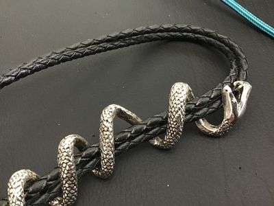 Sale Photo Thumbnail #255: Unique western bolo tie featuring a silver-tone coiling snake slide piece wrapped around a braided black leather/synthetic cord with silver-tone tips. Shows detailed scale texturing and open mouth snake design. Good condition with light wear.