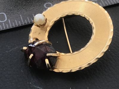 Sale Photo Thumbnail #58: Vintage circular gold-tone brooch featuring textured, engine-turned design details, accented with a single faux pearl and a prong-set faceted deep purple stone/glass element. Includes a standard pin mechanism on the reverse. Shows minor surface wear consi