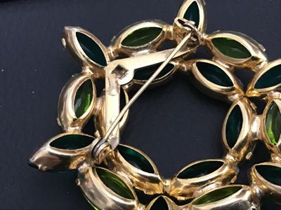 Sale Photo Thumbnail #212: A vintage gold-tone wreath brooch set with alternating emerald green and peridot lime green navette/marquise cut rhinestones in prong settings. Features a rollover safety clasp mechanism. Measures approximately 2 inches in diameter. Good vintage condition