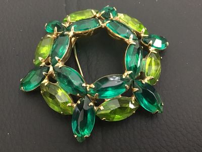 Sale Photo Thumbnail #208: A vintage gold-tone wreath brooch set with alternating emerald green and peridot lime green navette/marquise cut rhinestones in prong settings. Features a rollover safety clasp mechanism. Measures approximately 2 inches in diameter. Good vintage condition