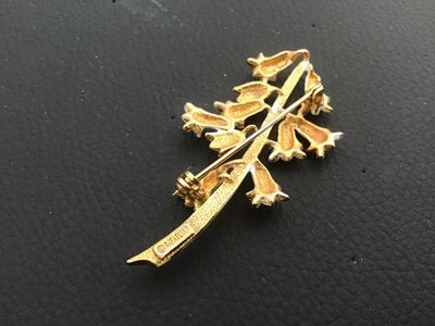 Sale Photo Thumbnail #19: Vintage Rafaelian floral pin featuring white enamel bell-shaped flowers on a green enamel stem with gold-tone backing and detail. Stamped '© RAFAELIAN' on the reverse. Features a standard pin back with a rotating clasp mechanism. Shows minor signs of vin